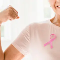 Lächelnde Frau mit rosa Schleife zeigt Bizeps. Onkologische Physiotherapie stärkt den Körper und gibt Hoffnung.