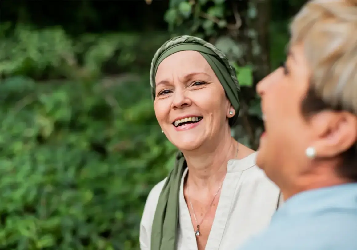 Zwei Frauen lachen im Freien. Die vordere Frau trägt ein Kopftuch und wirkt durch psychoonkologische Beratung gestärkt.