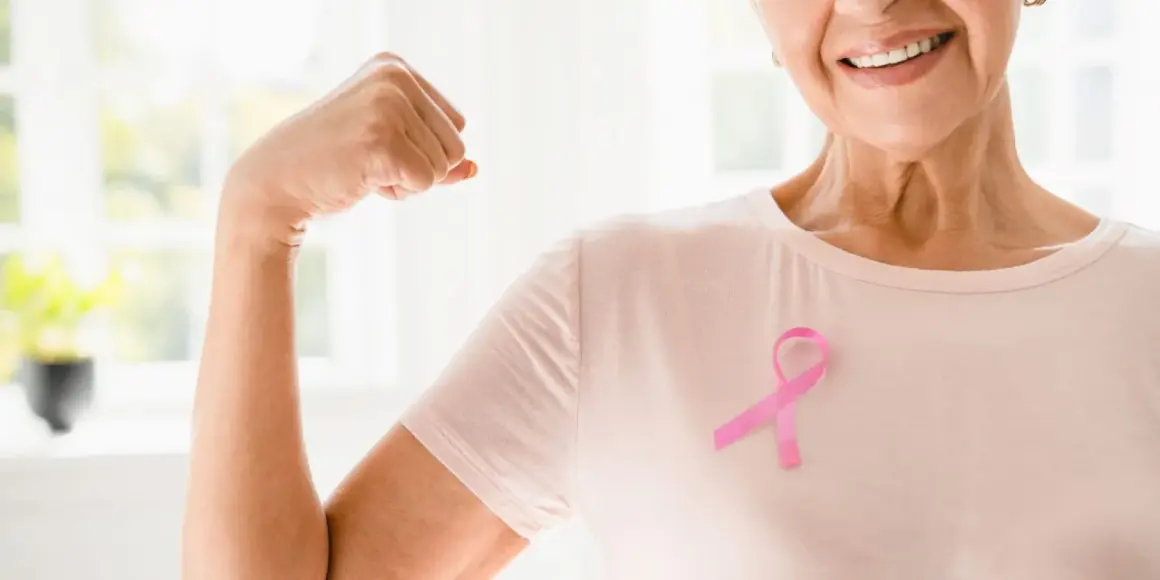 Lächelnde Frau mit rosa Schleife zeigt Bizeps. Onkologische Physiotherapie stärkt den Körper und gibt Hoffnung.