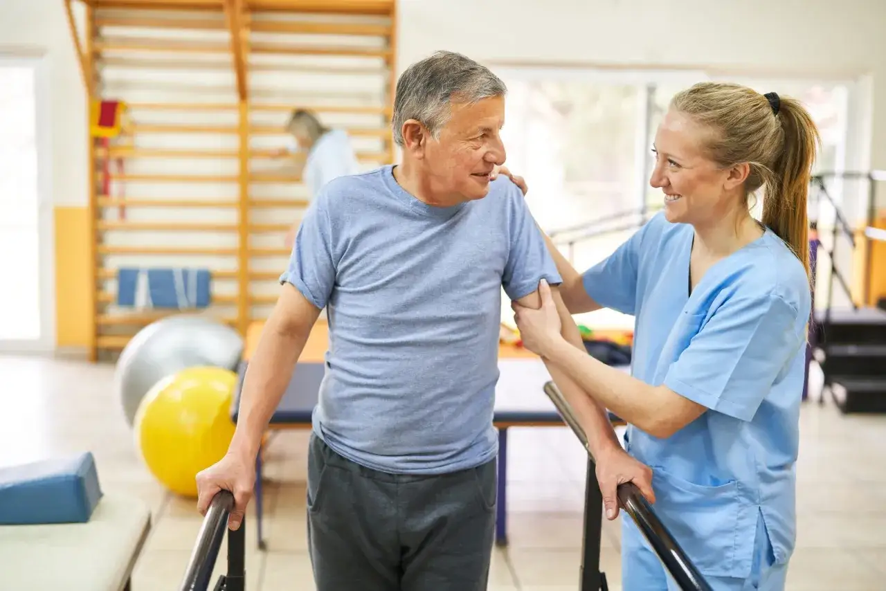 Ein älterer Mann übt mit einer Physiotherapeutin im Rahmen der onkologischen Physiotherapie das Gehen an einer Stange.