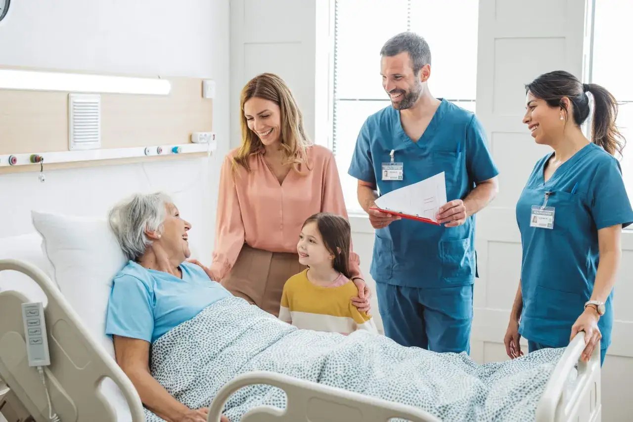 Ärzte und Familie bei der onkologischen Pflegevisite. Eine Patientin im Bett lächelt, während ihre Tochter und die Eltern zuhören.