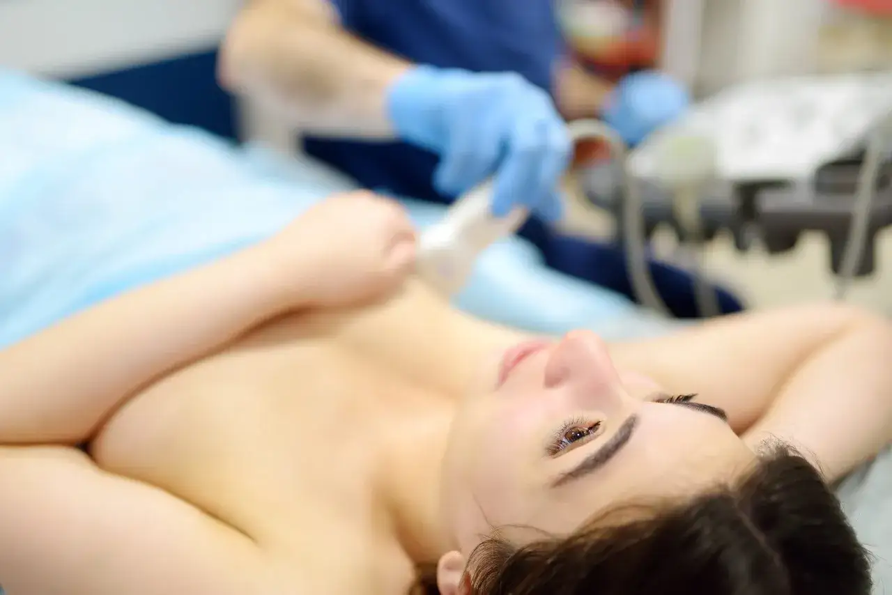 Eine Frau liegt auf einer Liege, während ein Arzt mit blauen Handschuhen eine Knochenszintigraphie durchführt. Sie wirkt angespannt, vielleicht wegen Platzangst.