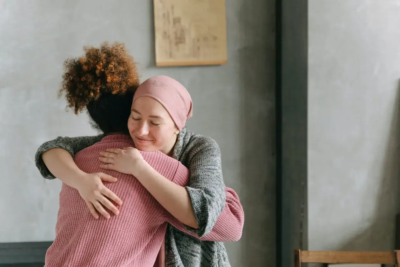 Zwei Frauen umarmen sich. Eine trägt eine rosa Kopfbedeckung nach Chemo, die sie vielleicht selbst gemacht hat.